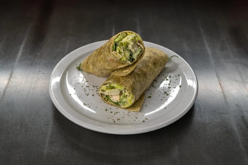Two halves of a chicken Caesar wrap, filled with lettuce and dressing, on a white plate.